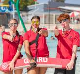Three lifeguards point at the camera holding lifesaving equipment in an effort to recruit new lifeguards.
